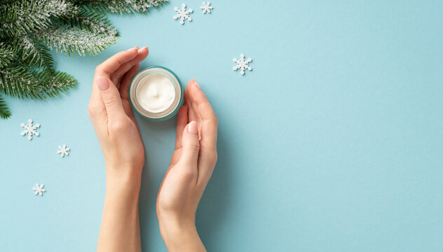 Winter Season Skin Care Concept. First Person Top View Photo Of Young Woman's Hands Small Jar Of Cream Snowflakes And Spruce Branches In Hoarfrost On Isolated Pastel Blue Background With Copyspace