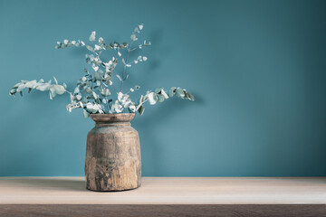 Rustic vase with dry eucalyptus leaves on wooden table against blue green wall. Home decor.