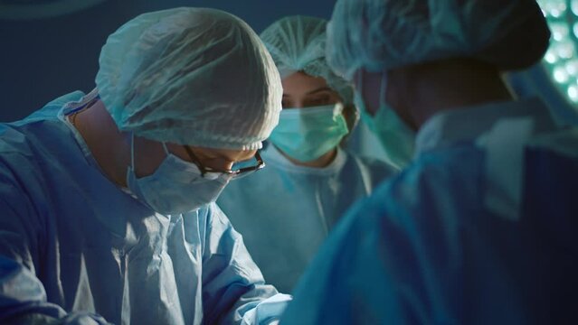 A Male Doctor Wearing A Full Surgeons Uniform Is Performing Intense Surgery While His Two Assistants On Either Side Of Him Are Helping Him Out