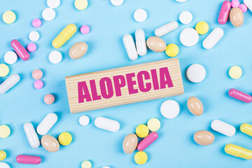 On a blue background, multi-colored pills and a wooden block with the text ALOPECIA. View from above. Medical concept