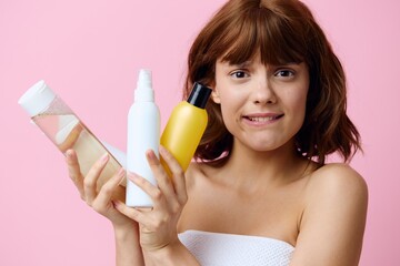 a sad, preoccupied woman stands on a pink background, wrapped in a white towel and holding a set of jars with facial care cosmetics in her hands, thoughtfully biting her lower lip