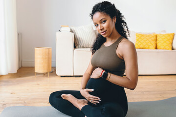 Pretty african american pregnant woman sitting on mat rubbing her belly, having rest after prenatal...