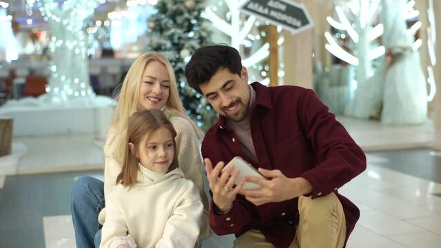 Happy young Caucasian family posing together for selfie picture using smartphone shopping in mall spending weekend together, standing smiling on background of festive Christmas decoration, slow motion