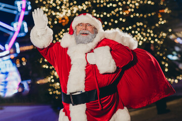 Portrait of positive cheerful grandfather santa carry presents bag arm hand waving congratulate tree garland lights evening outdoors