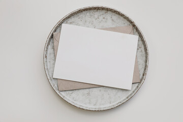 Neutral greeting card mockup. Blank paper invitation, craft envelope on ceramic plate isolated on...