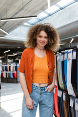 curly woman standing with hand in pocket and smiling at camera in textile shop - obrazy, fototapety, plakaty