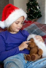 A cute little girl in a Santa hat is playing and hugging a teddy bear while sitting on the sofa in the living room. The concept of family holidays and traditions, love and care