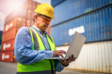 Engineer or foreman holding laptop and wears PPE checking container storage with cargo container background at sunset. Logistics global import or export shipping industrial concept.