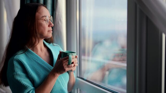 A middle-aged beauty woman with a cup at the window of the house. A happy woman in a bathrobe enjoys drinking hot coffee in the morning.
