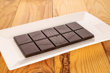 One bar of dark dark chocolate in a white ceramic plate on a wooden table, macro.