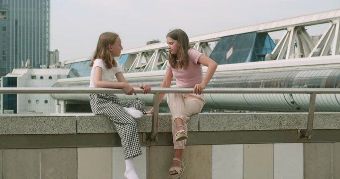 Female friends wearing casuals talking while sitting on surrounding wall in city during summer. Girls sits marble parapet and communicate together
