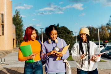 three latin college student girls walking together while talking and checking their mobile phones. student girls carrying folders, notebooks and smartphones.