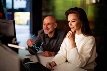  Group of casual business people sitting at table with computers and analyzing business information while working in office in late evening. Teamwork.
