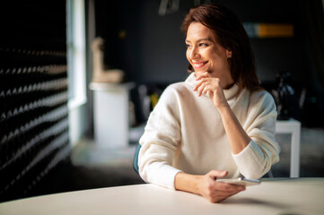 Portrait of attractive middle aged woman sitting at desk and looking away. Cheerful business woman wearing white sweater while working at office