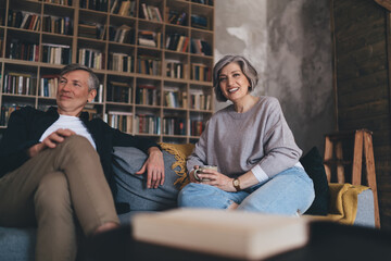 Fototapeta premium Cheerful aged man and woman sitting on couch against bookcase