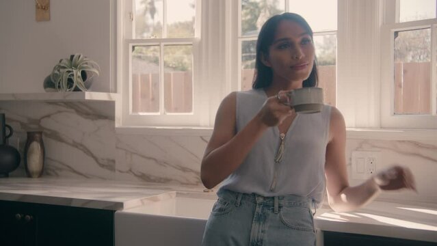 woman leaning on counter and drinking coffee or tea by a sunny kitchen window