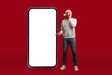 Holiday Sales. Surprised Arab Man Wearing Santa Hat Pointing At Blank Smartphone