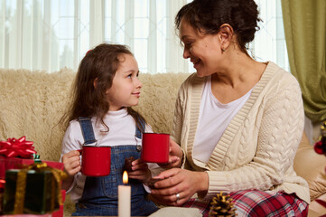 Beautiful multi-ethnic woman, happy loving mother and her lovely little daughter, enjoy time together by delicious hot chocolate drink at Christmas holidays. Xmas presents and lit candle on the table