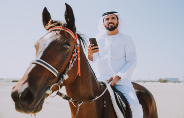 Young adult with Kandura, the emirates traditional clothes, riding his horse in the desert