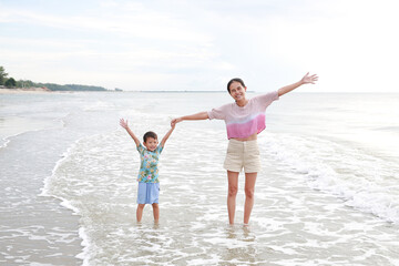 Asian mother and little boy standing at tropical sand beach with waving hands and open wide. Happy family mom and son having fun in summer holiday.