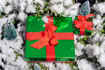 Emerald Christmas gift boxes with red bows next to small fir trees on green thuja branches covered with white fluffy snow, minimalist composition for Christmas and New Year in nature. flat lay