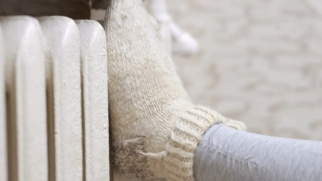 Woman puts feet in warm knitted socks on heating battery on blurred background. Housewife tries to get warm on cold winter day at home closeup