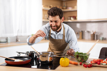 Cheerful adult european bearded man chef in apron prepares dinner for family, salts soup in modern kitchen - Powered by Adobe