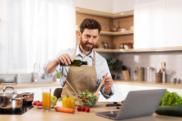 Fototapeta na wymiar Cheerful adult european bearded man in apron pours oil at salad in modern kitchen interior
