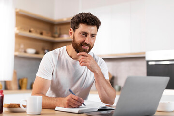 Happy handsome adult european bearded man making notes, watching video lesson on laptop