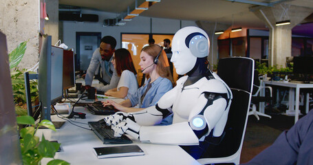 Robot working at computer among people. Maschine typing on keyboard in office. IT team of future. Futuristic worker. Humanoid work at call center. Support job. Selling concept. Technologies. - obrazy, fototapety, plakaty