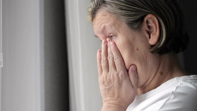 Depressed mature woman cries standing near window in bedroom. Female person wipes tears feeling heartache after loosing loved relative closeup