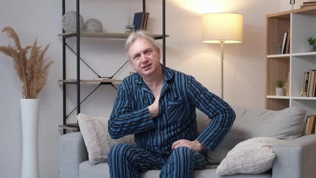 Heart attack. Aged man. Health problem. Shocked elder male in pajamas suffering from pain grasping chest sitting sofa breathing hard light room interior.