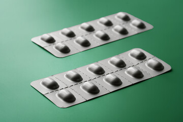 Two gray opaque metallized blisters with medical pills or capsules on a green gradient background. Selective focus