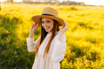 Medium shot portrait of pretty positive ginger young woman wearing straw hat and white dress standing posing on beautiful field of green grass looking at camera, in sunset sunlight on summer evening.