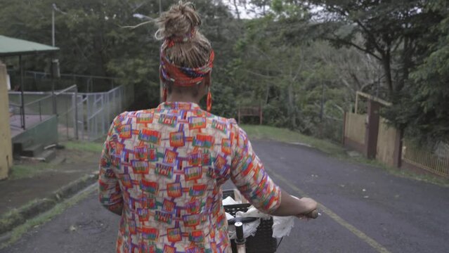 Mujer afro caribe&ntilde;a irreconocible caminando a espaldas con una bicicleta al lado rodando por la calle 