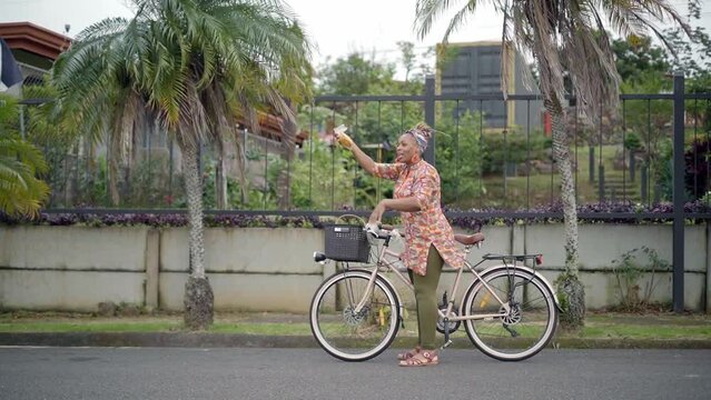 Imagen en video de una mujer afro caribeña de cuerpo completo con ropa colorida vendiendo en la calle sus productos en una bicicleta muy feliz
