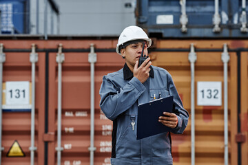 Waist up portrait of black woman wearing hardhat and talking to radio while working at shipping docks with containers