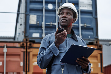 Waist up portrait of black male worker wearing hardhat and talking to radio in shipping docks with...