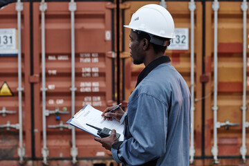 Side view portrait of young male worker wearing writing on clipboard in shipping docks with containers, copy space