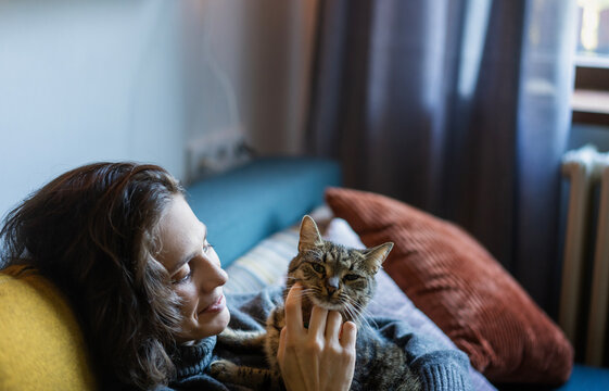 Cheerful smiling young woman in a warm sweater resting at home with a gray cat in her arms