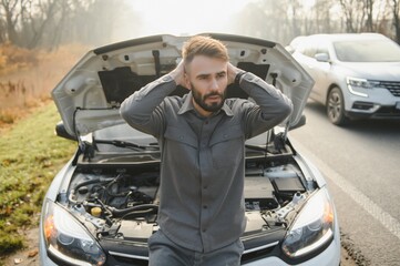 Fototapeta na wymiar Young upset casual man trying to fix his broken car outdoors. Man waiting for towing service for help car accident on the road. Roadside assistance concept.