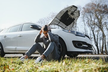 Picture of frustrated man sitting next to broken car with open hood