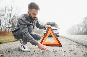 A young man near a broken car with an open hood on the roadside.