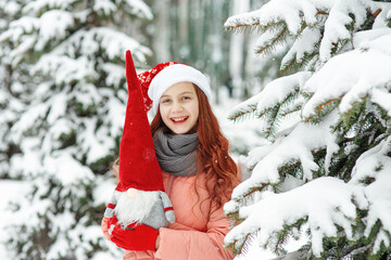 Young merry joyful laughing girl teenager in red hat standing among the snow-covered firs, holding...