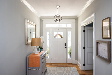 An open large and wide interior front door hallway foyer with transom, hanging light fixture, coastal colors and entry way table and wood floors - Powered by Adobe