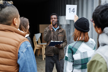 Back view of young woman holding number card bidding on container at outdoor auction in shipping...