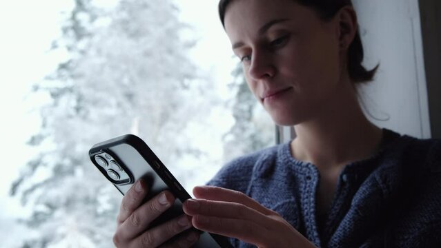 Selective focus of attractive young woman sitting on cozy windowsill and holding mobile phone enjoying beautiful winter nature. Smartphone application market, podcast listening. Relaxation concept