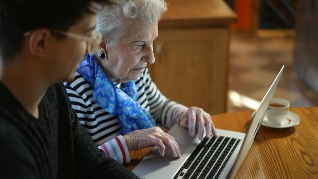 Closeup of elderly senior woman learning to use a laptop computer from teen male grandson with tea.