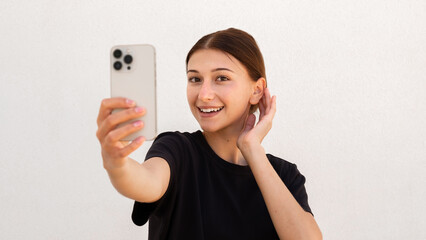 Portrait of happy young woman smarten up while looking at phone over white background. Caucasian woman wearing black T-shirt taking selfie or using mirror app. Mobile technology concept