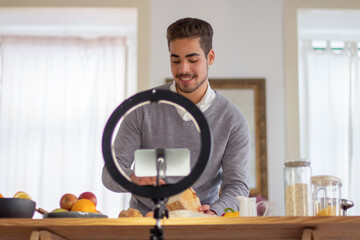 Fototapeta na wymiar Portrait of happy male culinary blogger vlogging online. Smiling man standing near table with food holding tasty bread talking about recipes of its cooking on camera. Healthy food, vlogging concept
